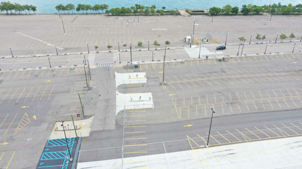 Color photograph of an empty surface parking lot adjacent to the Detroit Riverwalk and the Detroit River itself.