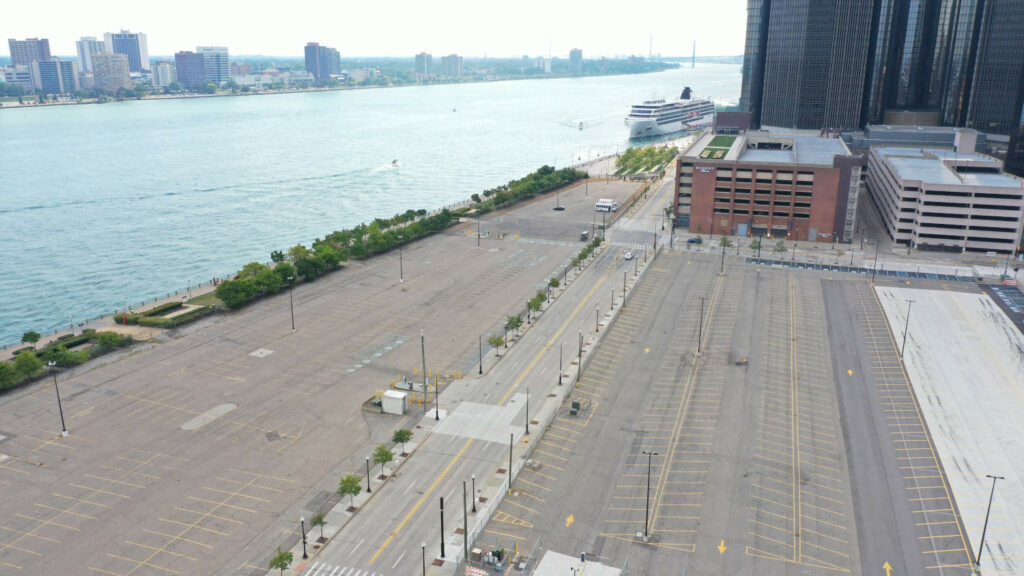 A color photograph showing several empty surface parking lots and two large parking structures near the Renaissance Center and adjacent to the Detroit Riverwalk and Detroit River.