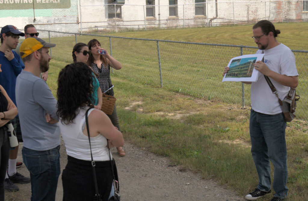 Walking Tour attendees gather around Francis Grunow of Detroiters for Parking Reform as he shows a photograph of the former Park Plaza Hotel at the corner of Second Avenue and Ledyard Street. Demolished ca. 2017. Now a vacant lot.